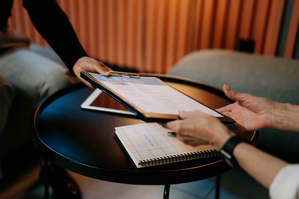 An individual hands another individual a clipboard with a document on it, as one might in a job interview