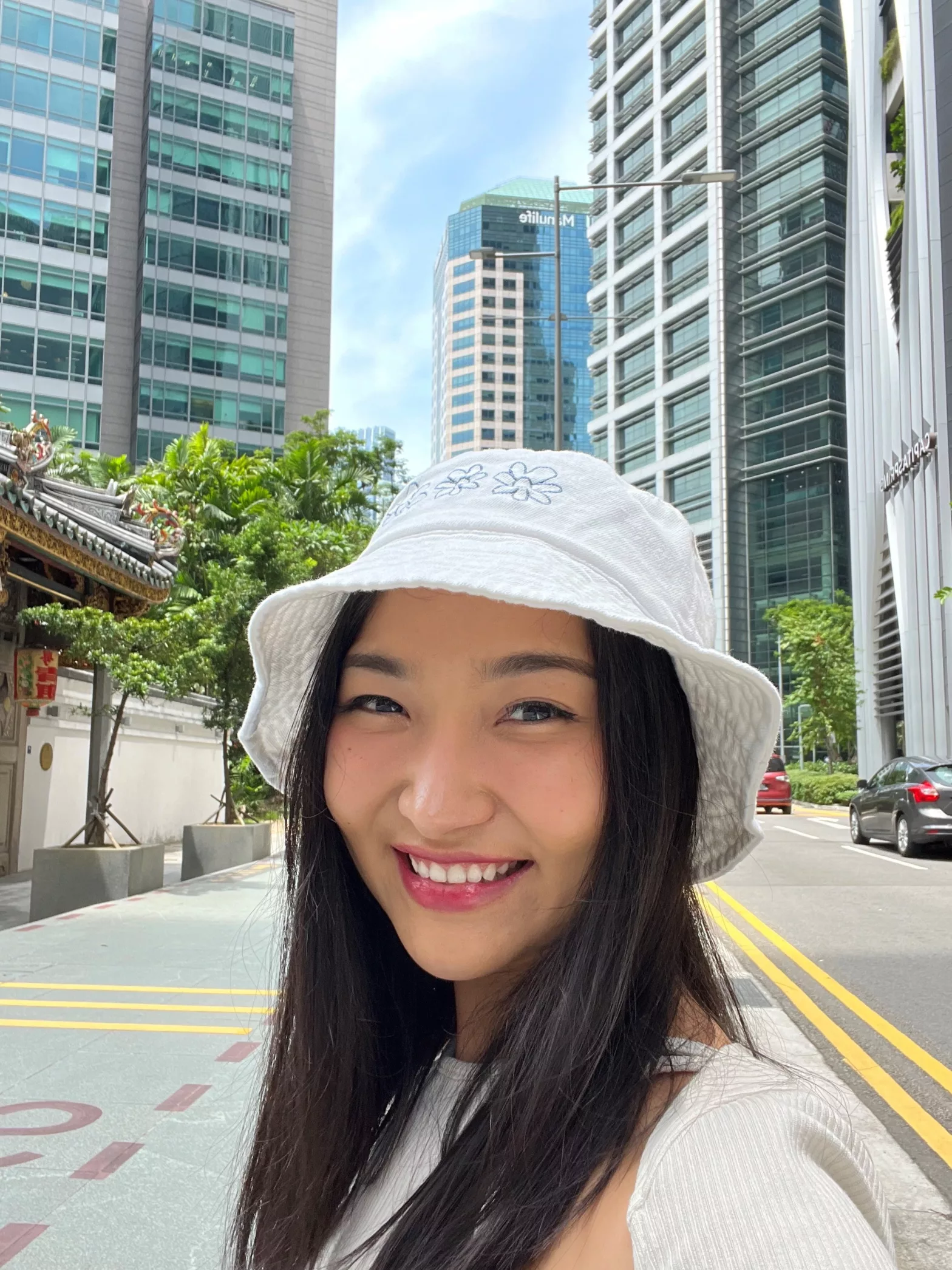 A young woman with black hair and a white hat stands in the middle of a city block with tall buildings in the background